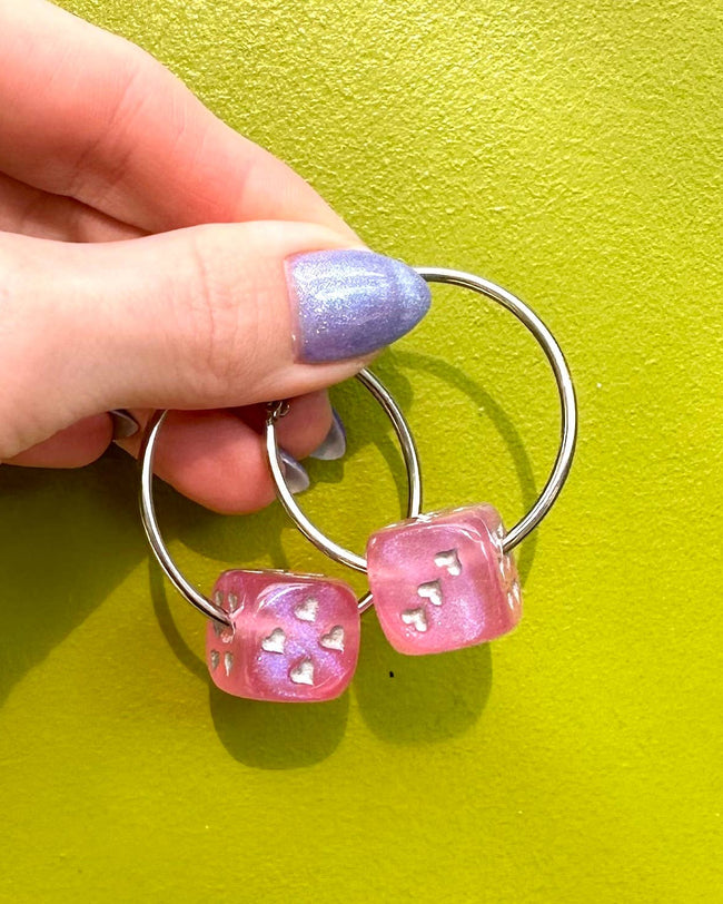 Hand holding a silver ring with pink dice-shaped charms against a green background
